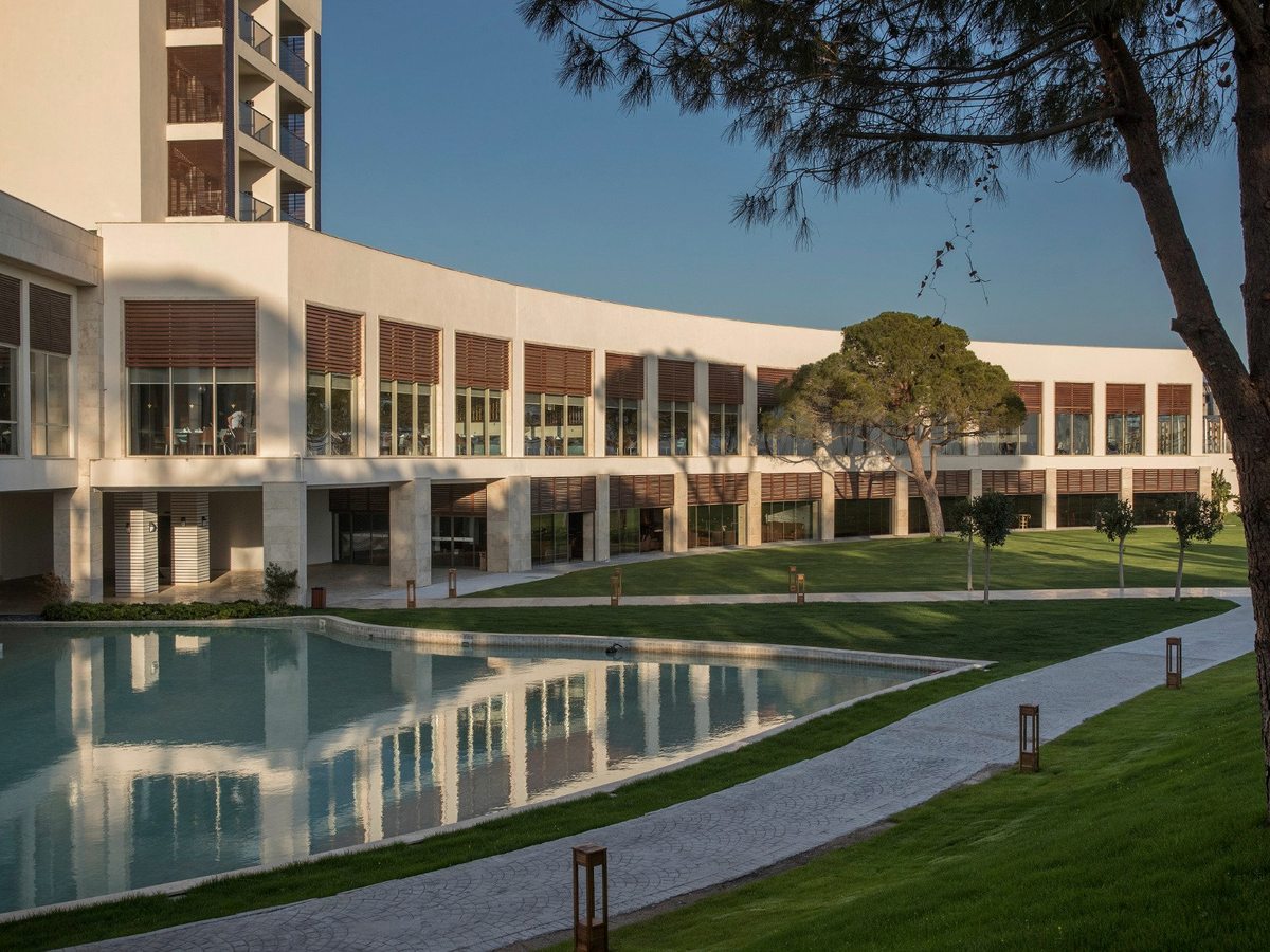 A long curved white hotel wing with rows of shuttered windows above lawns and an ornamental pool, with umbrella pines alongside.
