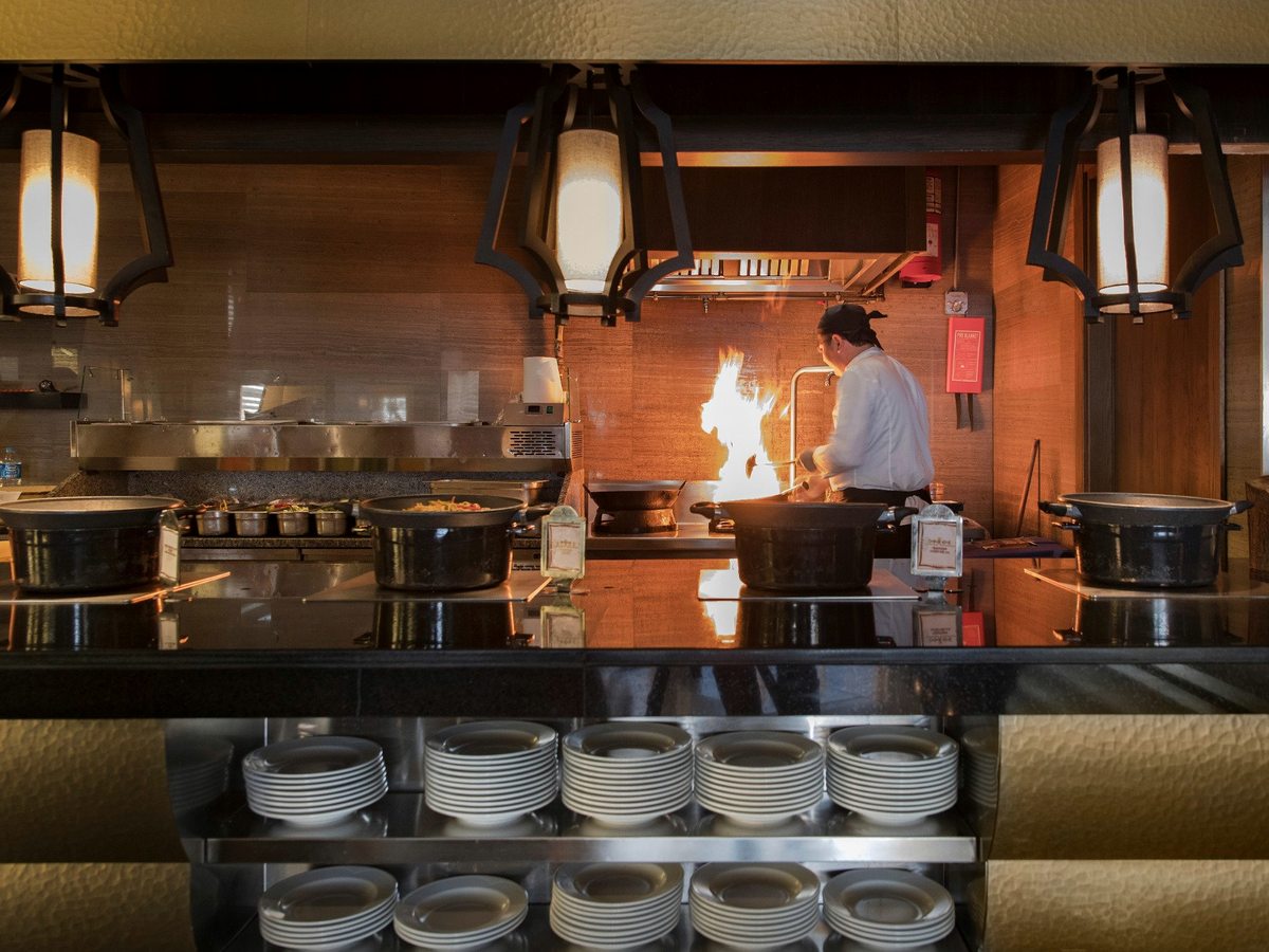 A chef cooking over a flame behind an open kitchen counter, with stacked plates on a shelf below the counter.