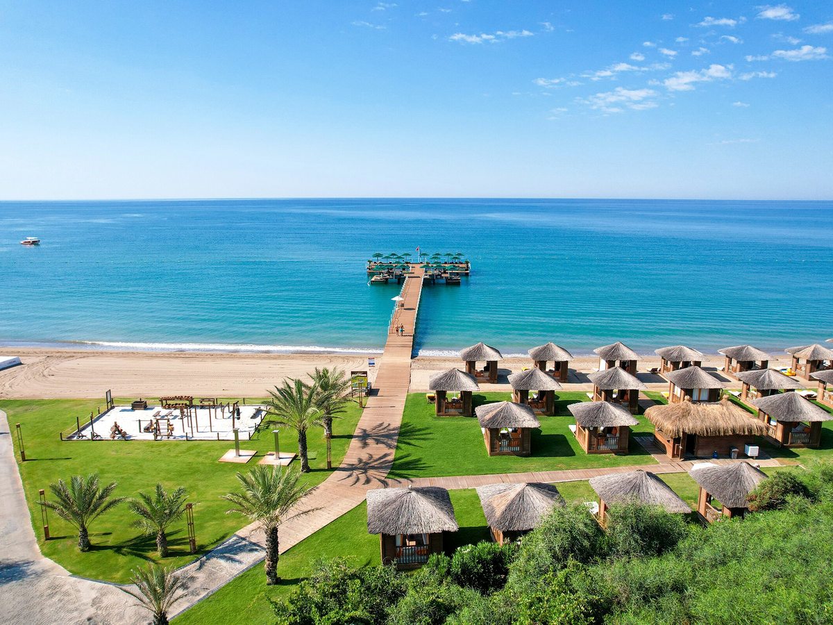 Rows of thatched timber cabanas on a lawn behind a sandy beach, with a long wooden pier running out into a calm blue sea.