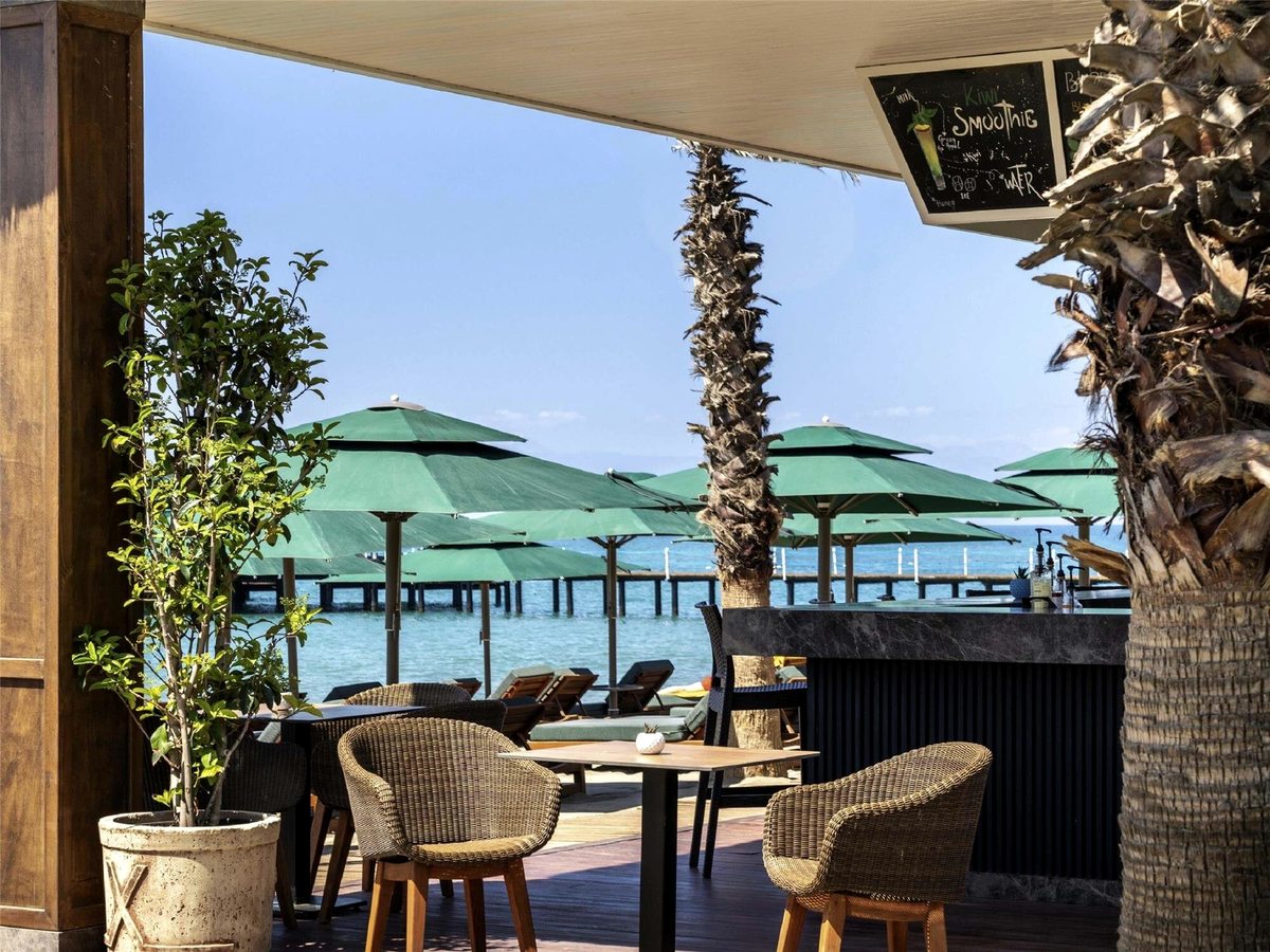 A shaded bar counter with wicker chairs and low tables on a timber deck, green parasols on the sand and the pier over the sea behind.