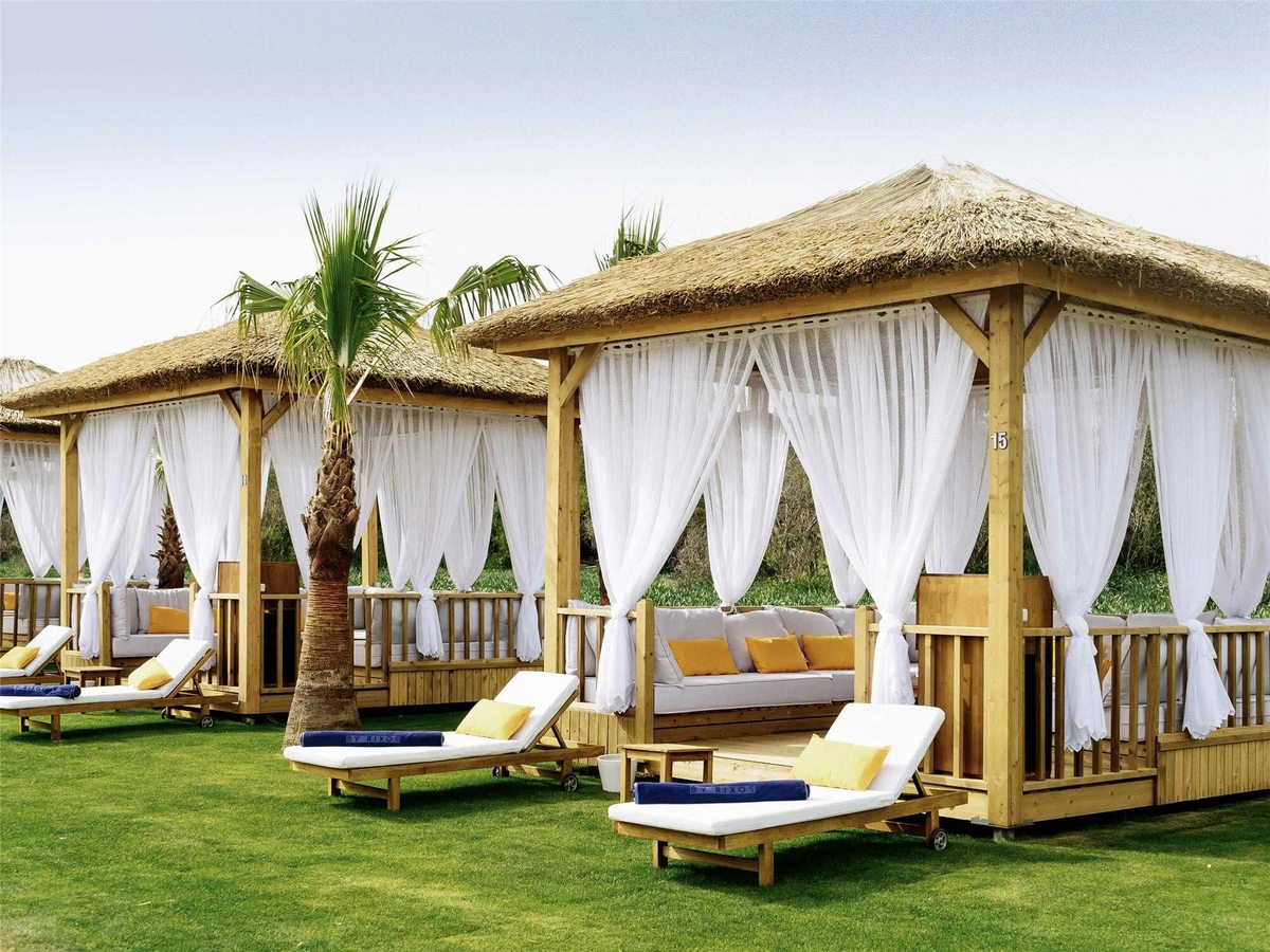 Thatched timber cabanas with white curtains and cushioned daybeds on a mown lawn beside a palm tree.