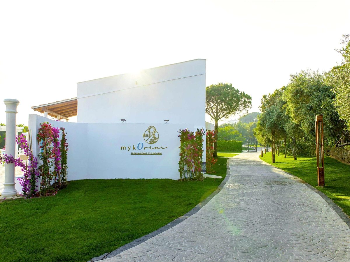 A white rendered wall carrying the myKorini name and logo, flanked by climbing bougainvillea, beside a paved path across a lawn.