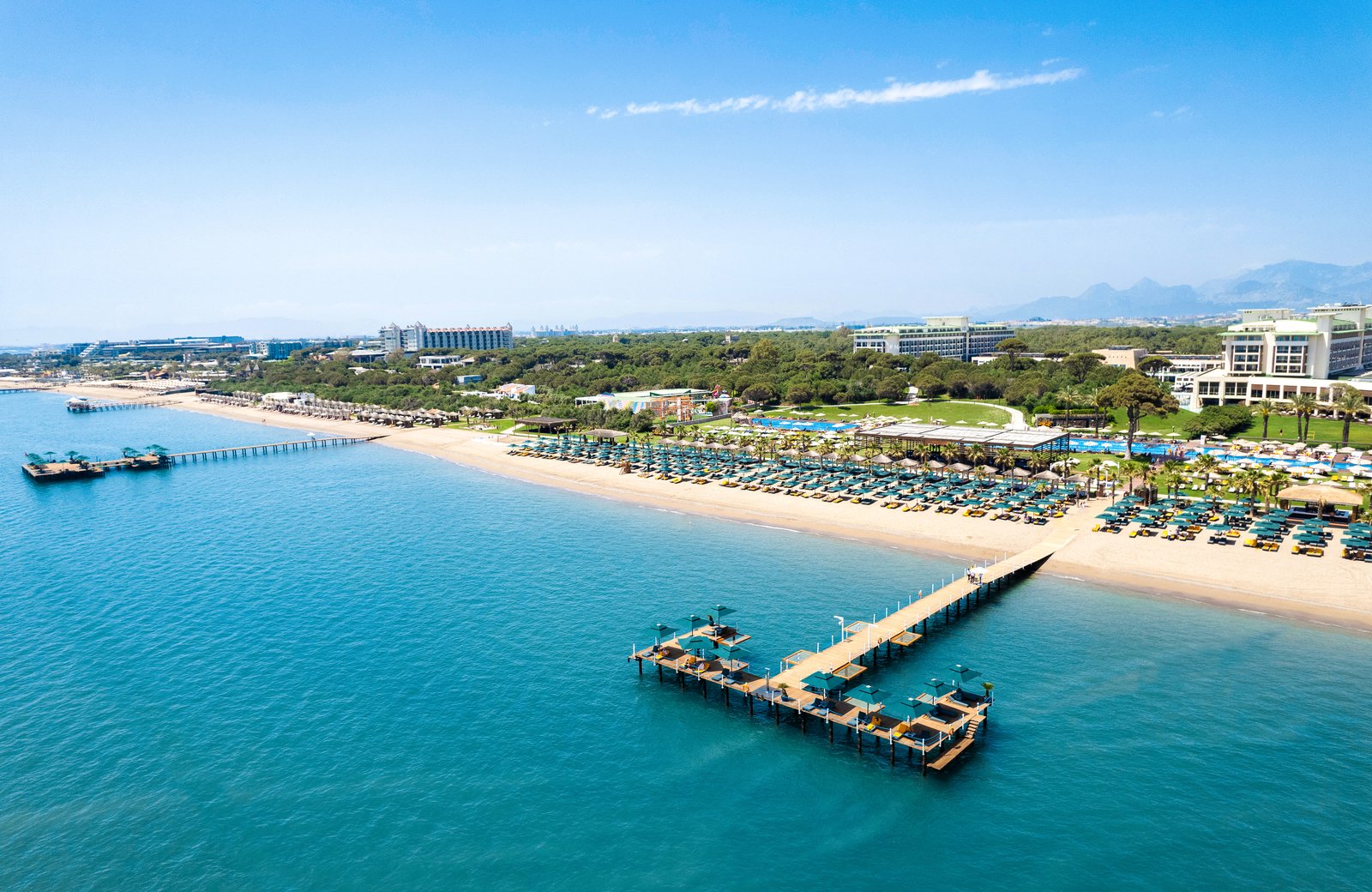 Rixos Premium Belek from the sea: a long sandy beach lined with rows of parasols and sunbeds, two wooden piers over turquoise water, and the resort's buildings among pine woodland behind.