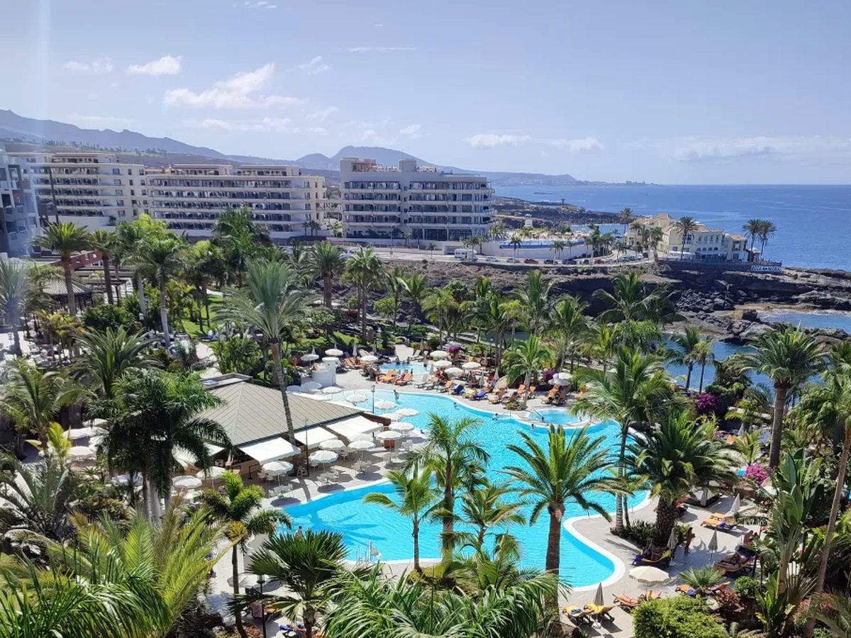 A high view over palm-filled hotel gardens and two large pools ringed with loungers, with a rocky shoreline, further hotels and mountains along the coast.