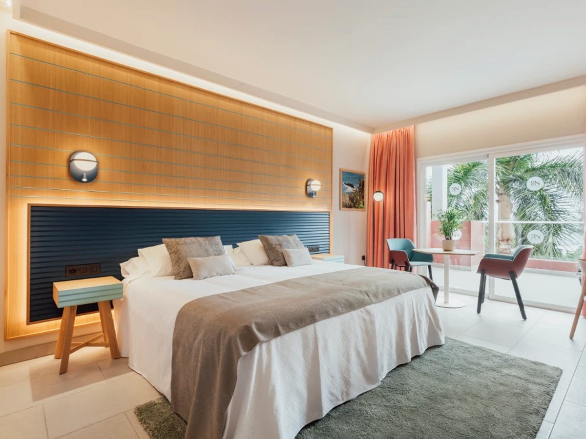 A hotel bedroom with a double bed against a wood-panelled and navy headboard, coral curtains, and a table and two chairs beside glass doors onto a balcony with a coral parapet and palms outside.
