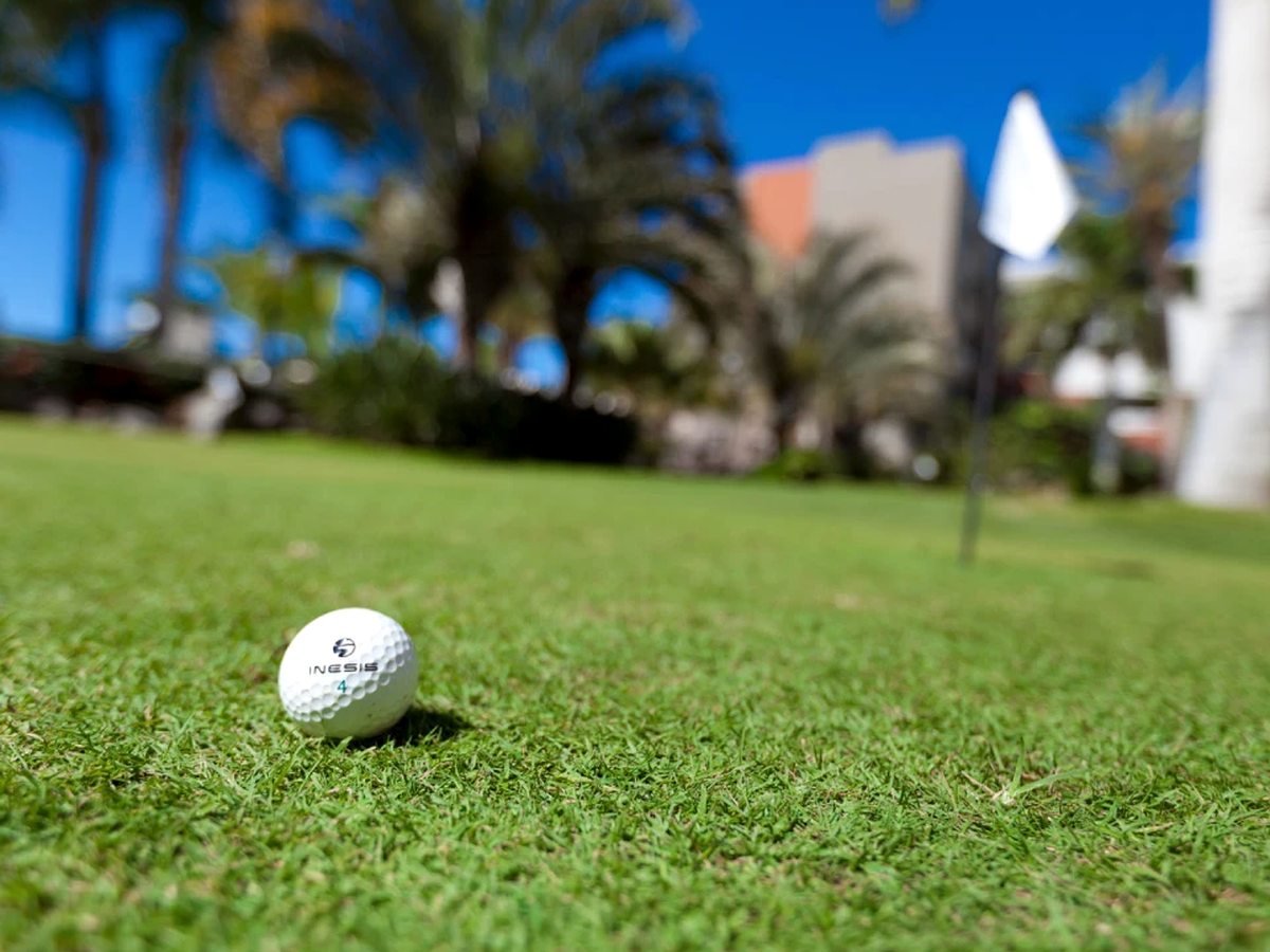 A white golf ball resting on a close-cropped putting green, with a small white flag, palm trees and a hotel building blurred behind.