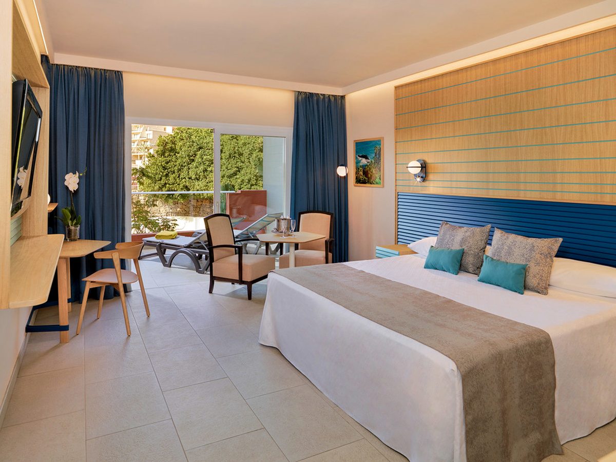 A bedroom with a double bed, blue curtains, a desk and wall-mounted television, and a balcony with a lounger and dining table looking onto trees.