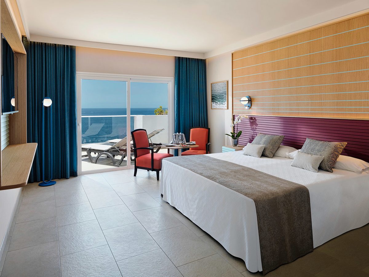A bedroom with a double bed, blue curtains and two red armchairs, and sliding doors onto a balcony with two sun loungers and open sea beyond.