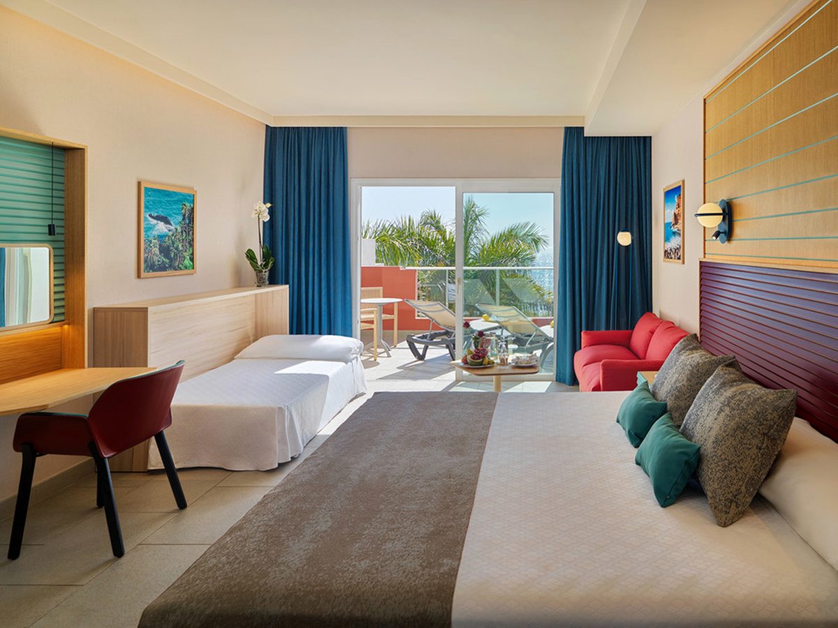 A room with a double bed, a made-up single bed alongside it, a red sofa, and glass doors onto a terrace with loungers and a table.