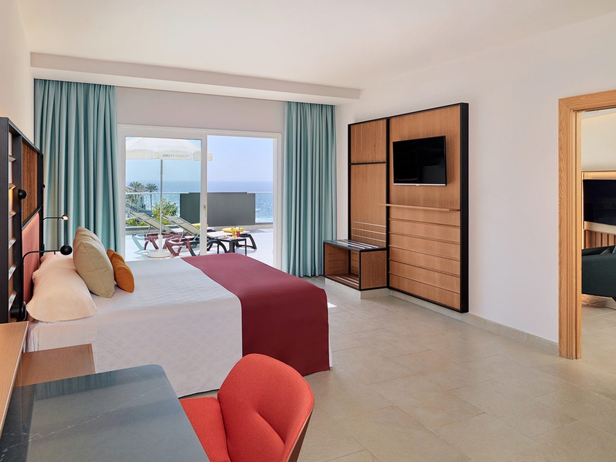 A bedroom with a double bed under a red runner, a wall-mounted television, a wide doorway through to an adjoining room, and glass doors onto a terrace with the sea beyond.