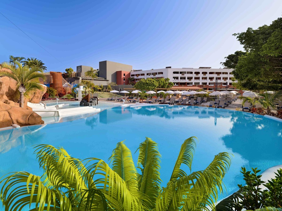 A large blue pool edged with sun loungers and white umbrellas, with a sandstone rock formation and white water slides at one end and hotel buildings behind.