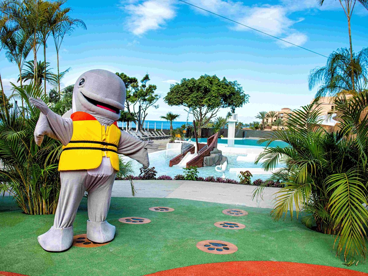 A costumed grey whale mascot in a yellow life jacket standing on a green play surface, with a pool, palm trees and the sea behind.