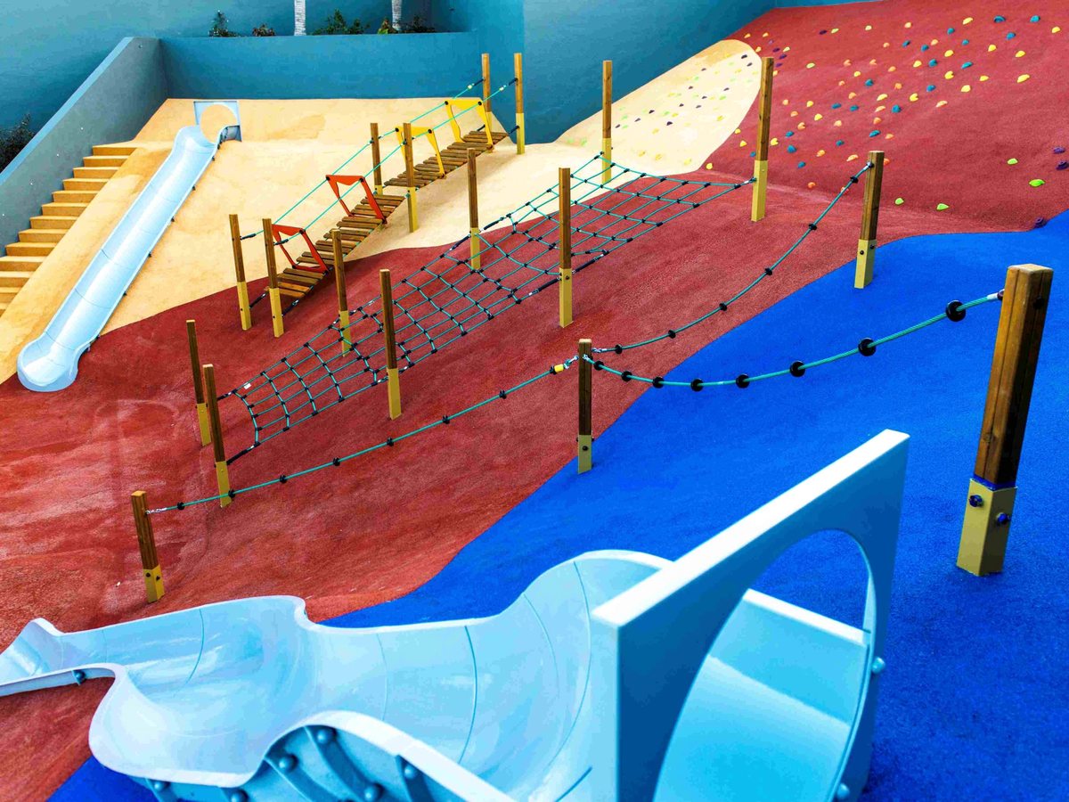 A play area with a red and blue rubber surface, wooden posts strung with rope nets, a suspension bridge, a climbing wall with coloured holds and two pale blue slides.