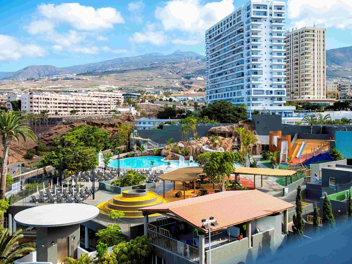 A hotel's water play area seen from above: a rock waterfall with white slides, a blue pool, a red and blue rubber play surface, palms, and apartment towers and mountains behind.