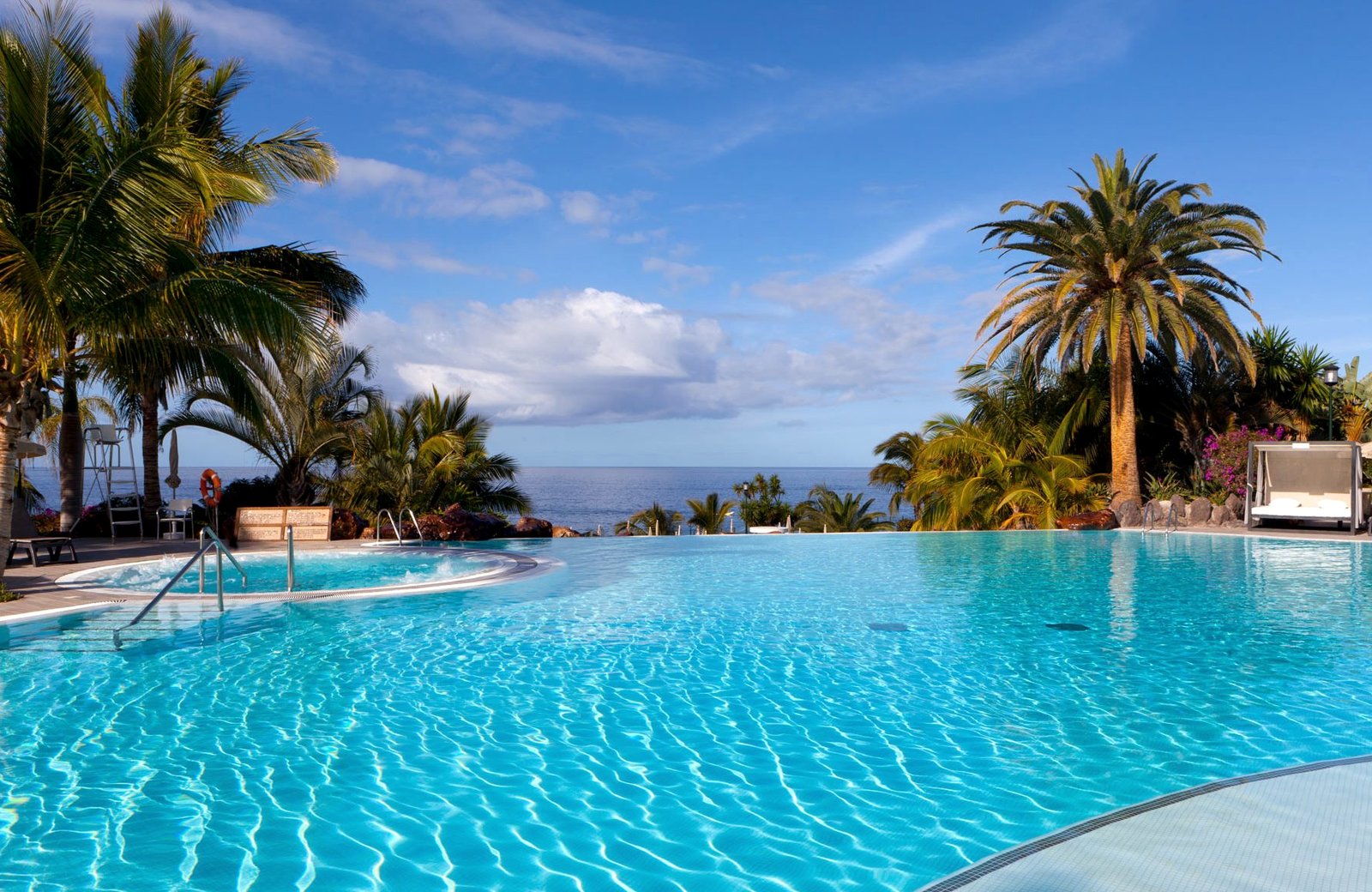 A wide infinity pool at Roca Nivaria Gran Hotel, palm trees on either side and the Atlantic on the horizon under a blue sky.
