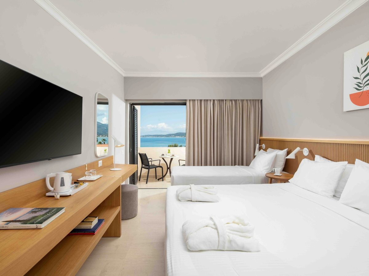 A bright room with a double bed and a single bed, a long pale-wood desk holding a kettle and tea tray, a wall-mounted television and open doors to a balcony with a table and chairs above the sea.