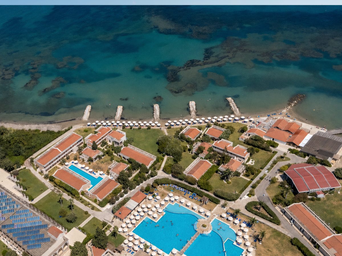 An aerial view of low red-roofed buildings and lawns with a long rectangular pool among them, and a line of white parasols along the sand at the shore.