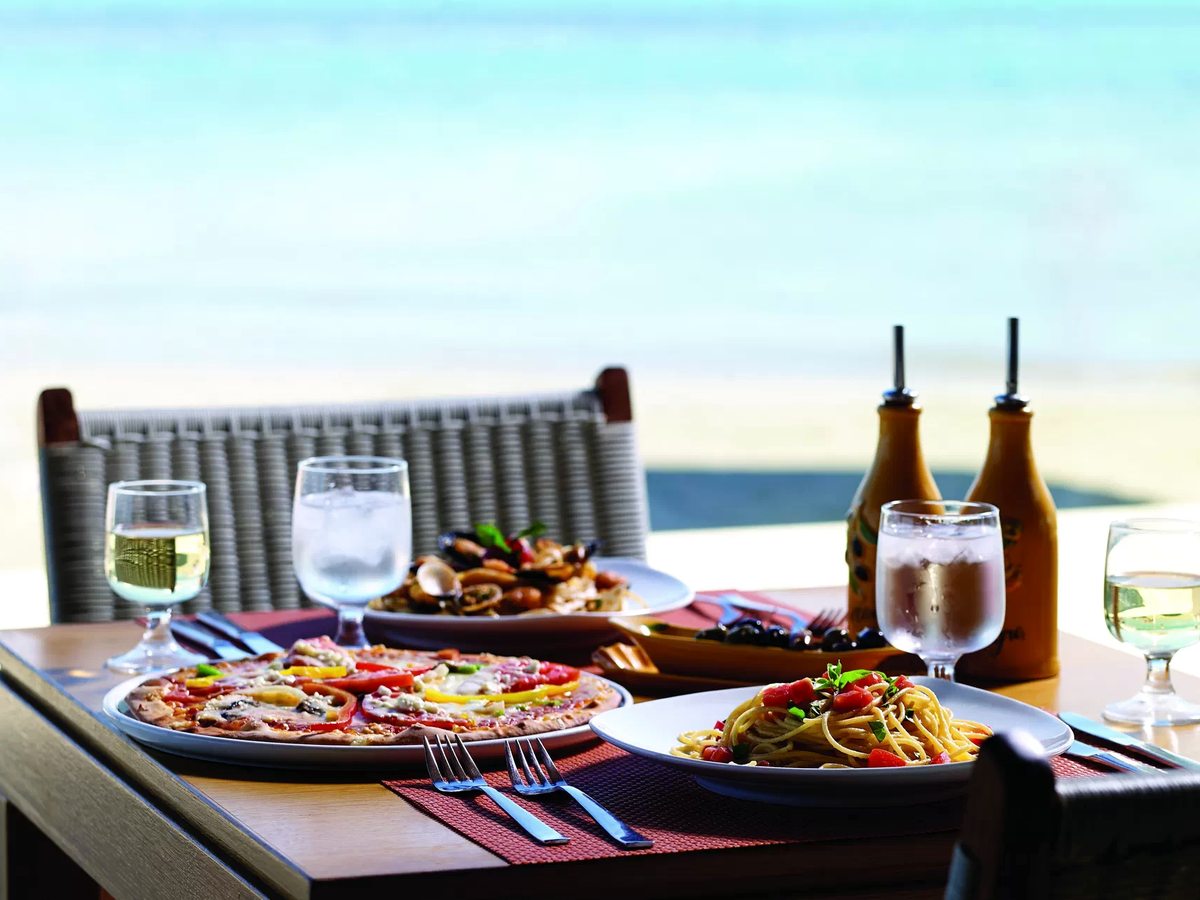 A table laid with a pizza, a plate of spaghetti, a dish of mussels and clams in their shells and glasses of white wine, with the sea behind.