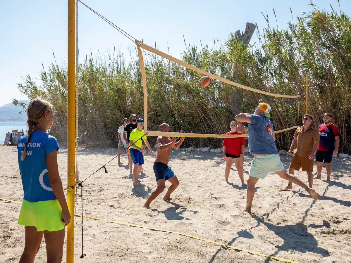 Guests playing volleyball on a sandy court with a yellow-taped net, a tall reed windbreak behind them and the sea to one side.