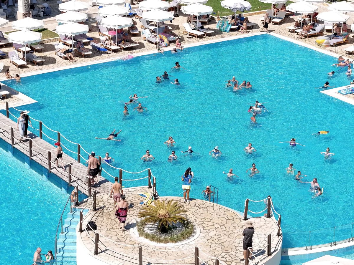 A large pool busy with swimmers, edged by rows of white parasols and sun loungers, with a planted stone island and a rope-railed walkway crossing the water.
