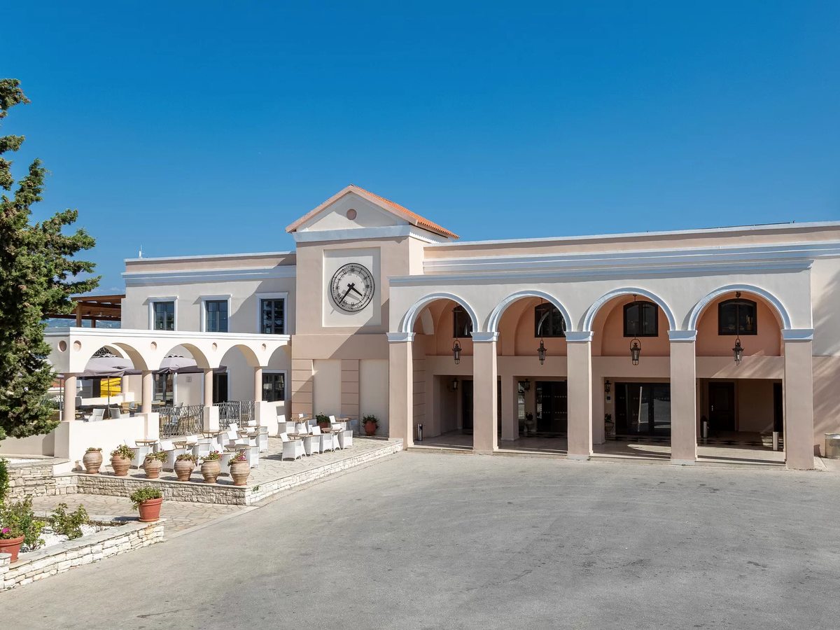 The cream main building in bright sun, with four tall arches over the entrance, a clock set into the gable above and a terrace of white chairs and potted plants to one side.