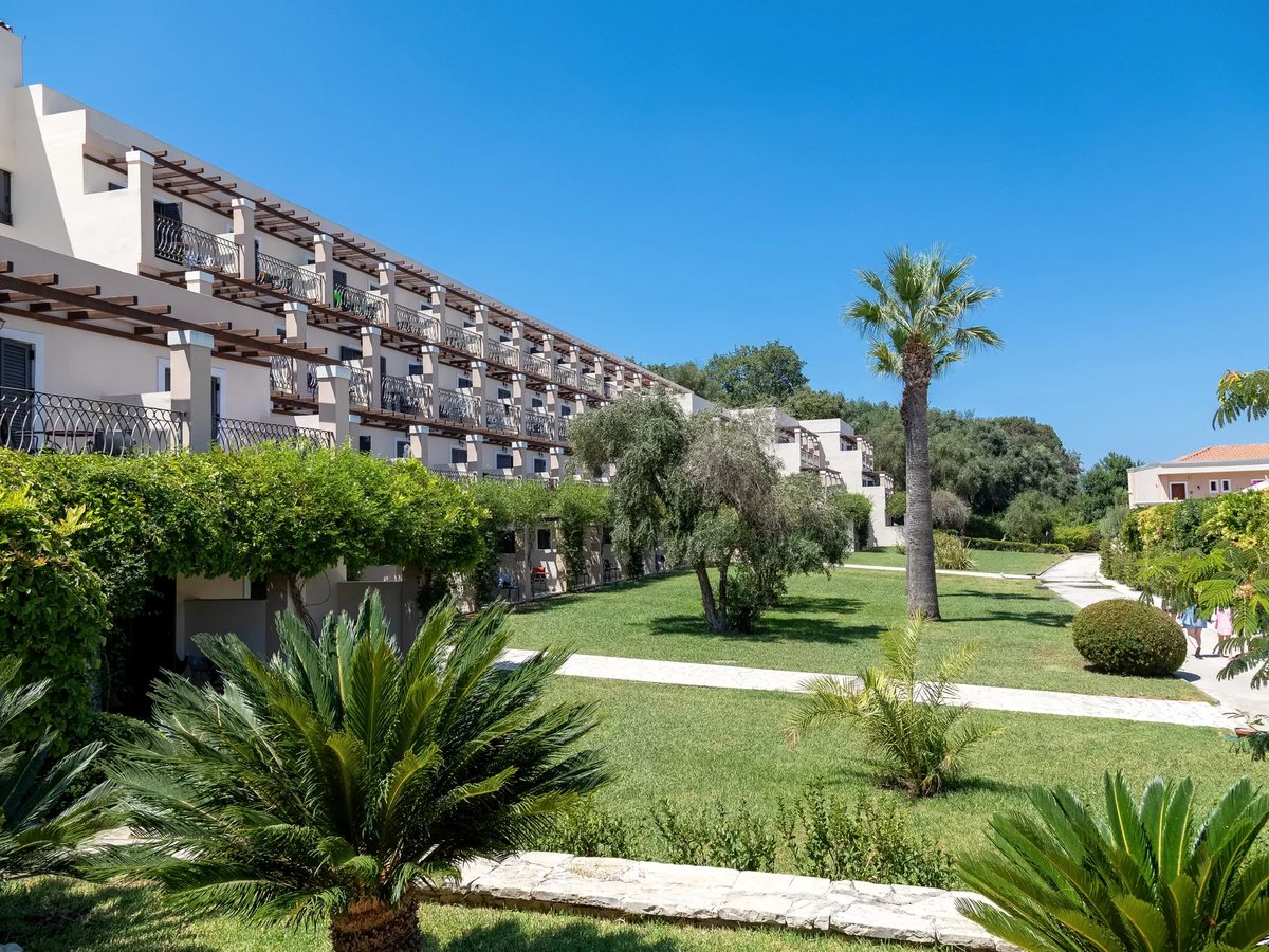 A long two-storey accommodation block with rows of balconies, seen across a lawn planted with palms and shrubs and crossed by a paved path.