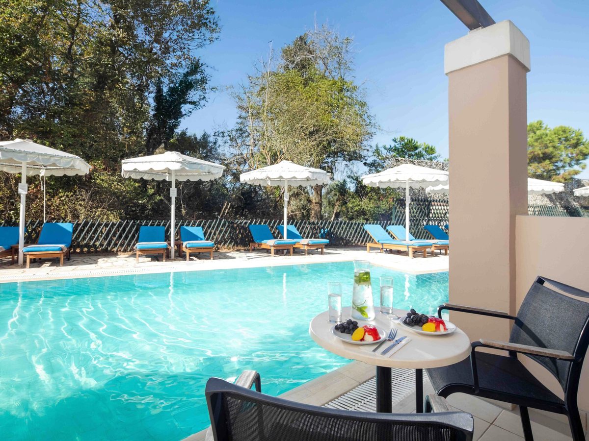 A pool seen from a shaded room terrace with a table set with fruit and water, blue loungers and white parasols lining the far side beneath trees.