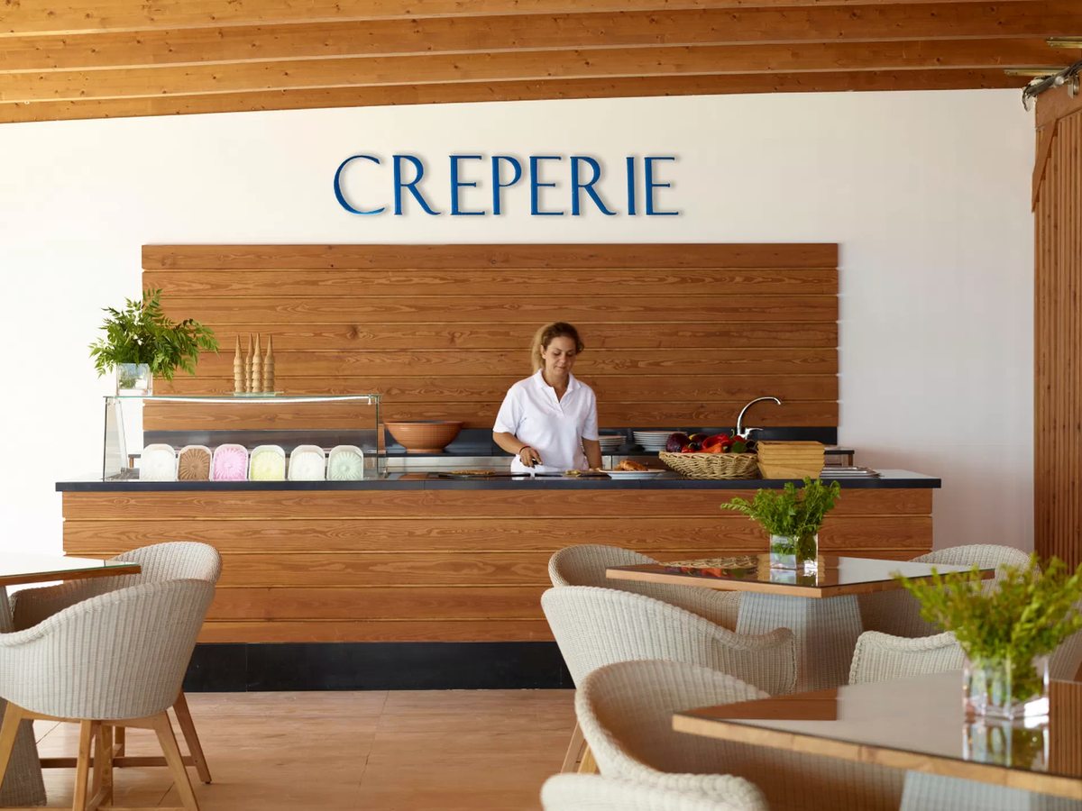 A timber-fronted counter beneath a sign reading Creperie, with tubs of ice cream in a chilled display, a member of staff working behind it and wicker armchairs at tables in front.