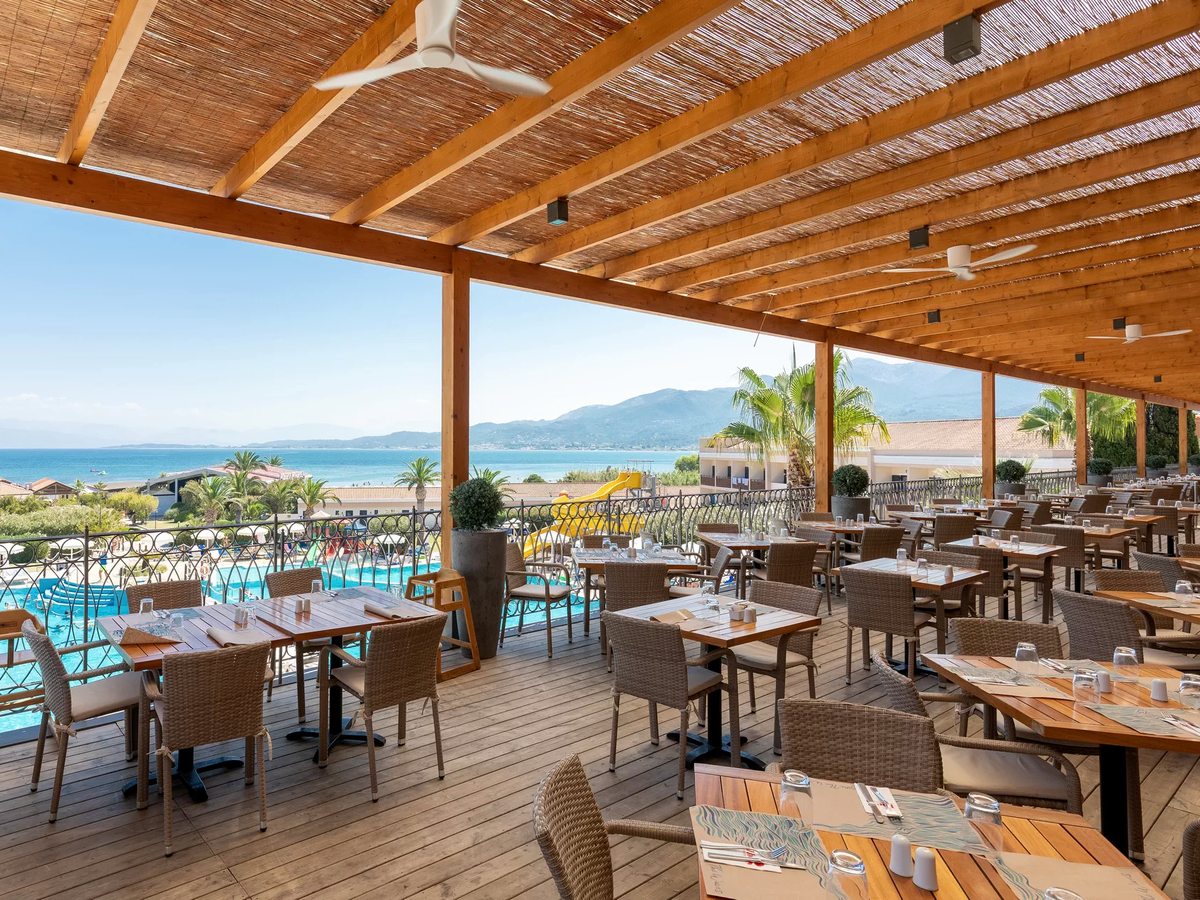 A large timber-framed terrace under reed shading, filled with laid wooden tables and wicker chairs, above a pool with yellow and blue waterslides and the sea and mountains beyond.