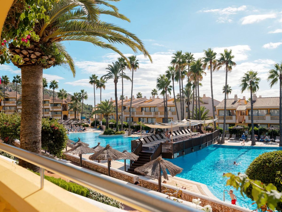 Low-rise yellow and cream apartment blocks with tiled roofs standing among tall palm trees around a large blue swimming pool edged with parasols and sun loungers.