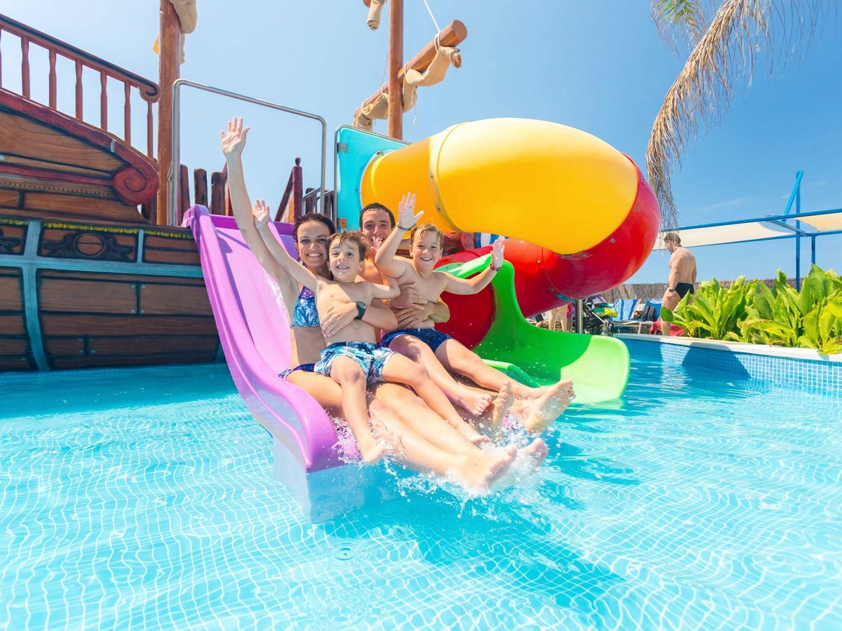 Children and adults coming down brightly coloured water slides into a shallow pool beside a wooden pirate ship structure.