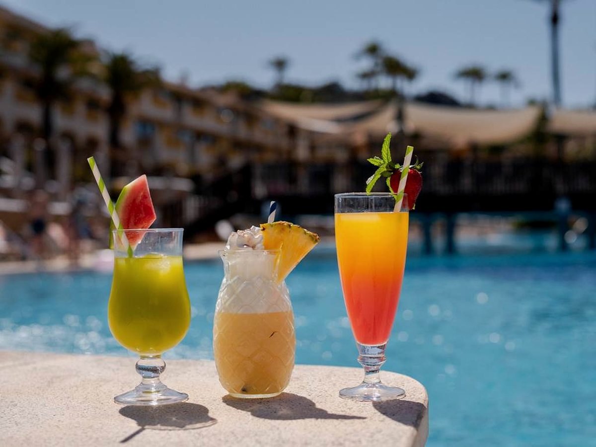 Three tall glasses of fruit drinks with straws and slices of fruit standing on a stone poolside ledge, with the blue pool and resort buildings behind.