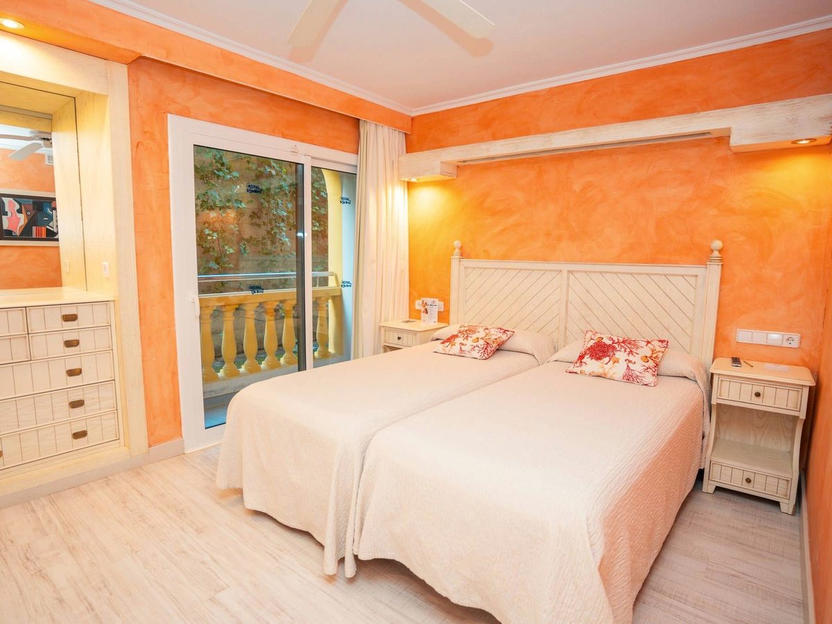 A bedroom with orange walls, two single beds pushed together under a pale wooden headboard, bedside tables, a chest of drawers and a balcony door.