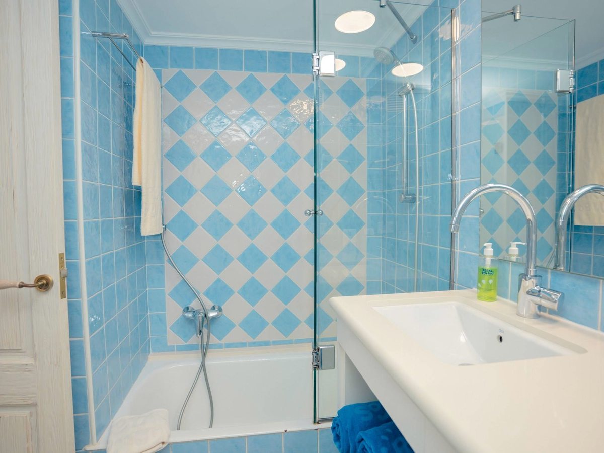 A bathroom tiled in blue and white with a bath under a glass shower screen, a white basin unit, folded towels and a toilet.