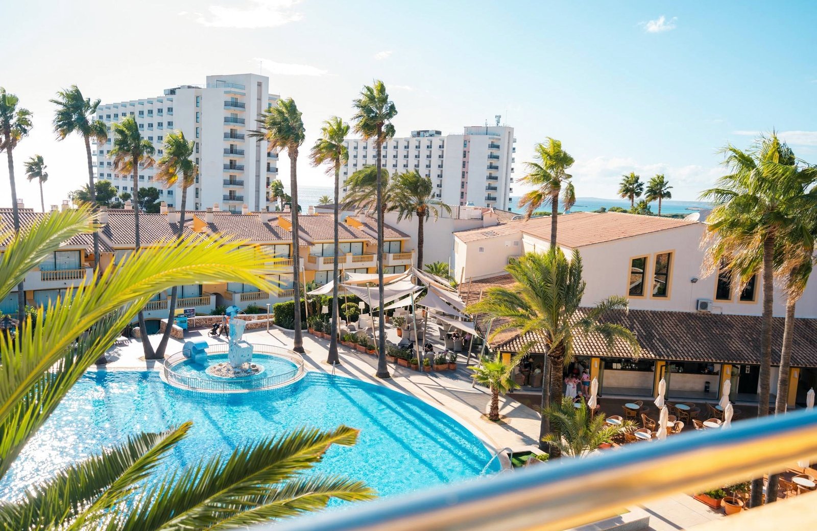 The pool and terrace at Royal Son Bou Family Club, ringed by palm trees and the resort's low-rise apartment blocks, with the sea beyond.