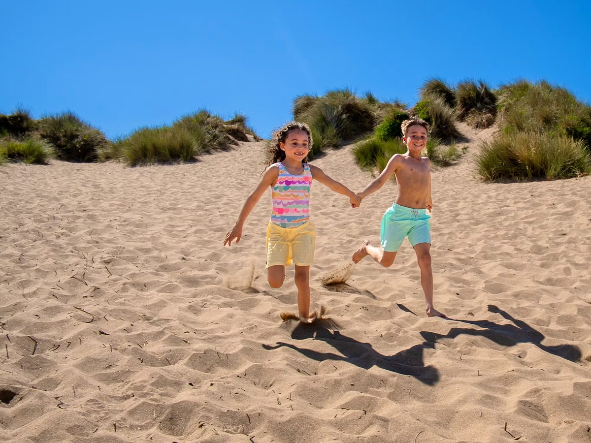 Two children running hand in hand down a slope of soft sand below marram-grass dunes under a deep blue sky.