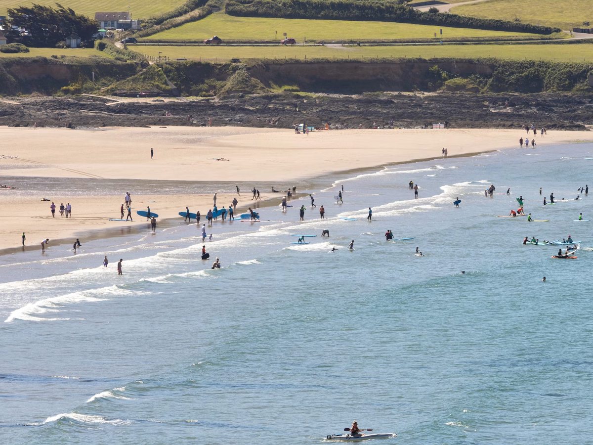 Dozens of surfers and swimmers in the breaking waves of a wide sandy bay, with a rocky shore and green fields on the land above.
