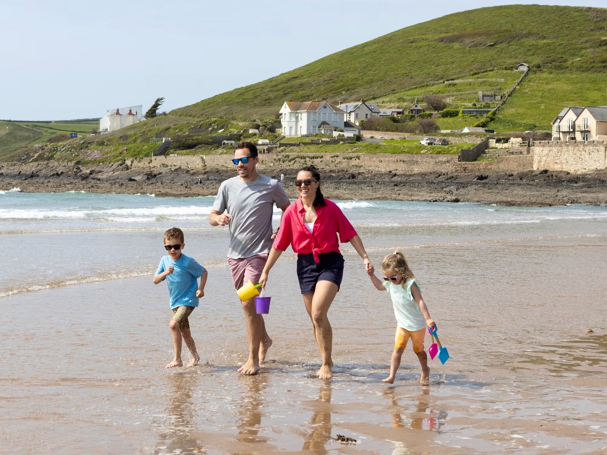 A family paddling through shallow water at the edge of a sandy bay, carrying buckets and spades, with a green headland and white houses behind.