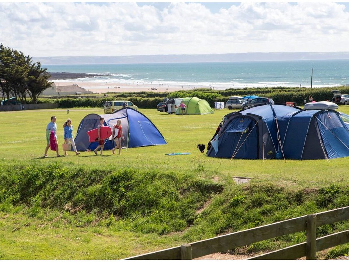 Family tents and campervans spread across a wide grass camping field, with a long sandy beach, breaking waves and open sea beyond the hedge.