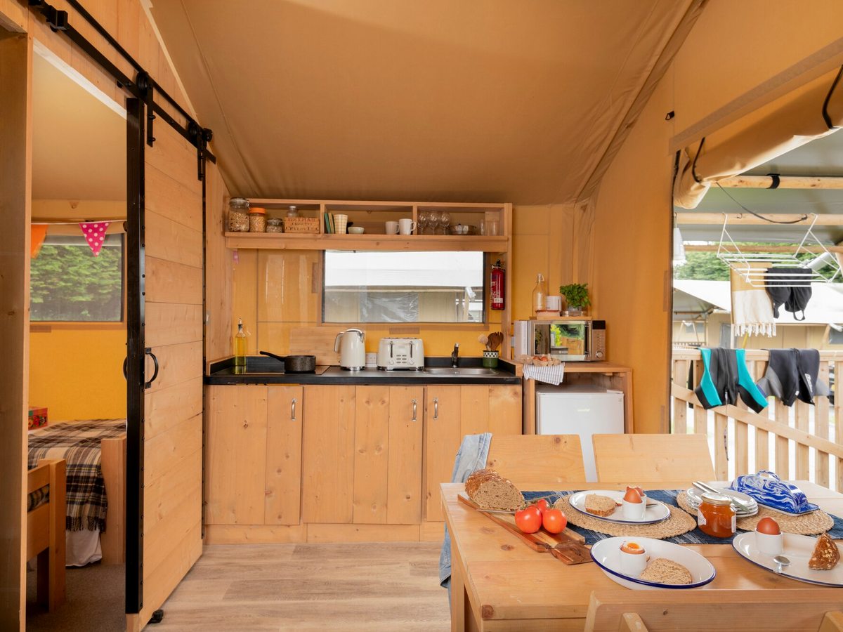 A safari tent kitchen with pine cabinets, a hob, kettle, toaster, microwave and fridge, a wooden table laid for breakfast and a bedroom through a sliding door.