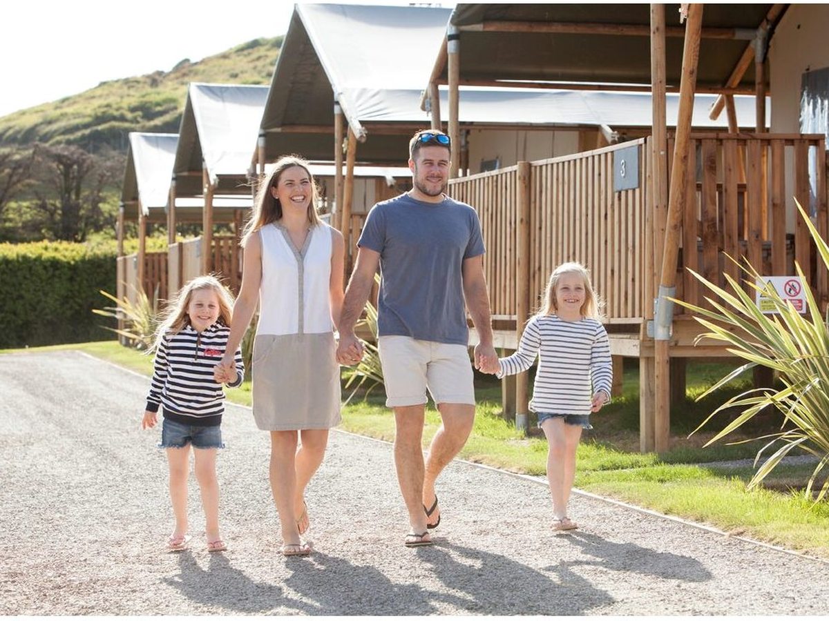 A family walking a gravel path past a row of numbered safari tents with canvas roofs and timber verandas, with a green headland behind.