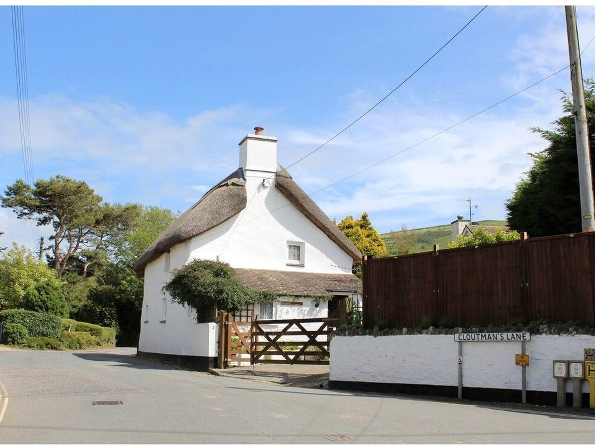 A whitewashed thatched cottage with a chimney and a wooden five-bar gate at a lane junction, with green hills behind.