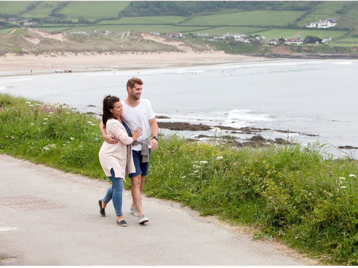 A couple walking a narrow tarmac path above a wide sandy bay backed by dunes, with green fields and houses on the far shore.