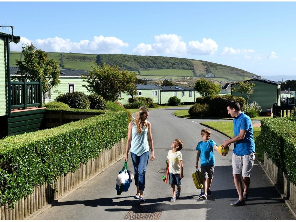 A family walking down a tarmac road between rows of green-clad static caravans behind clipped hedges, with a green hillside behind.