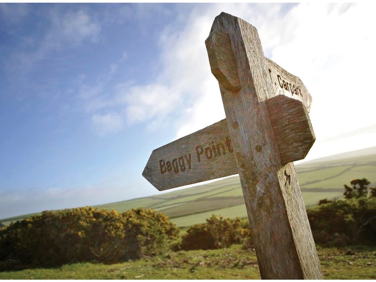 A weathered wooden fingerpost carved with Baggy Point and Car park, standing on a grassy headland with gorse in flower behind.