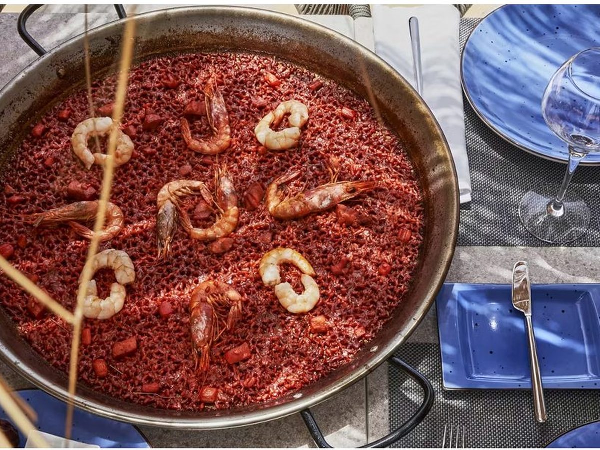 A wide steel pan of deep red rice studded with whole prawns and chunks of tomato, on a table with blue plates and a wine glass.