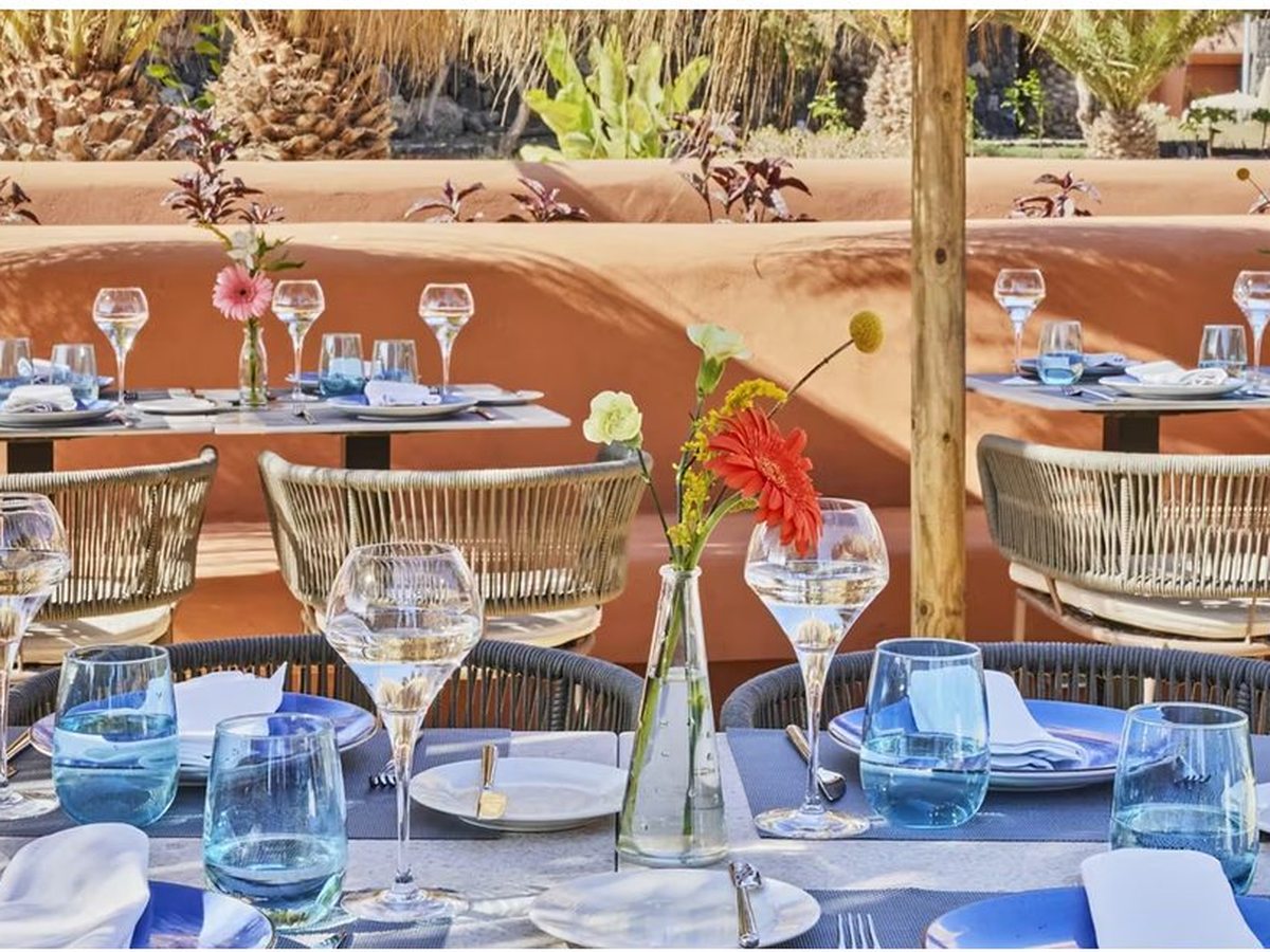 Outdoor tables laid with blue plates, blue glassware and small vases of flowers, set on a terrace against a curving terracotta wall with palms behind.