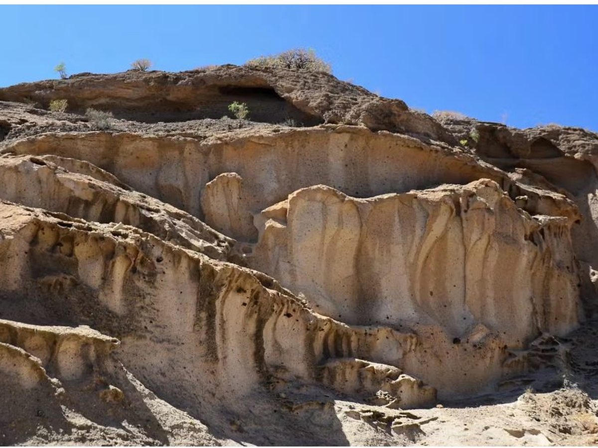 Steeply eroded pale volcanic rock faces, fluted and pocked with holes, with scrubby plants along the ridge under a deep blue sky.