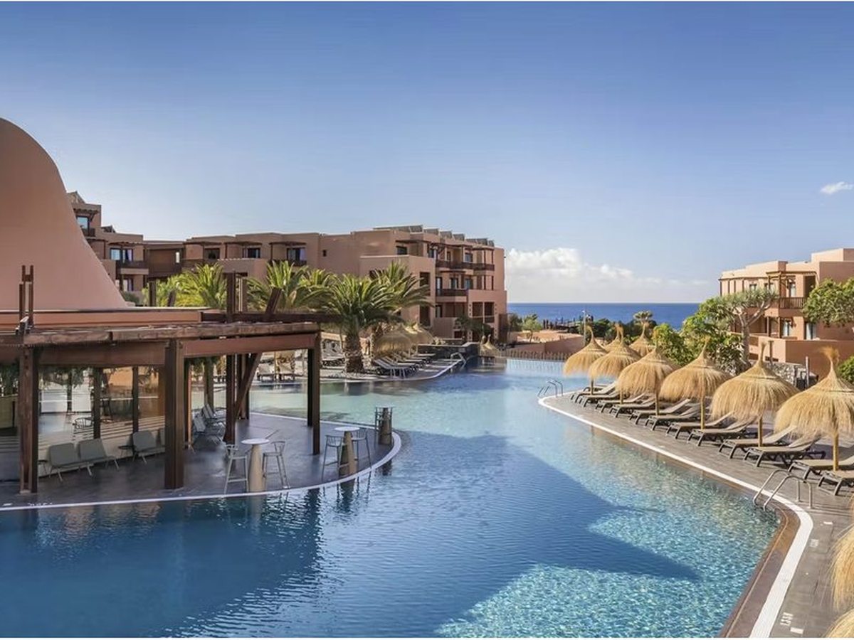 A wide curving lagoon-shaped pool with a timber pool bar pavilion at one edge, straw parasols and sun loungers along the other, and terracotta room blocks and the sea behind.