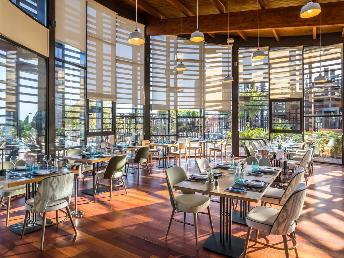 A large restaurant with a high timber ceiling and pendant lamps, pale patterned chairs at wooden tables laid with blue glassware, and full-height windows with roller blinds along both walls.